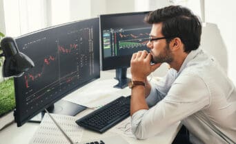 Successful trader. Back view of bearded stock market broker in eyeglasses analyzing data and graphs on multiple computer screens while sitting in modern office.