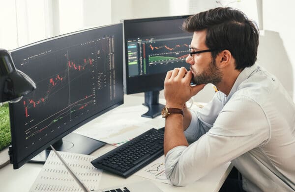 Successful trader. Back view of bearded stock market broker in eyeglasses analyzing data and graphs on multiple computer screens while sitting in modern office.