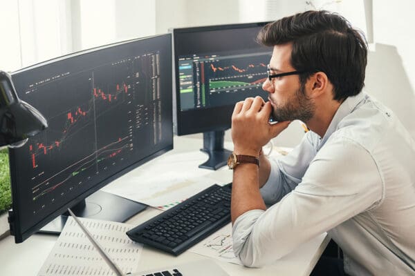 Successful trader. Back view of bearded stock market broker in eyeglasses analyzing data and graphs on multiple computer screens while sitting in modern office.